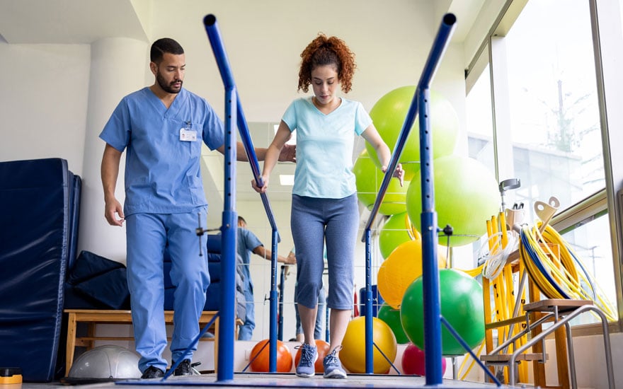 A young woman works with a physical therapist holding on to parallel bars.